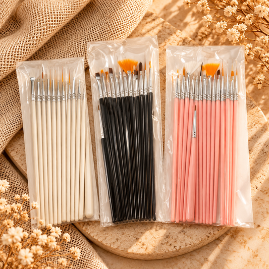 Three packs of paintbrushes arranged on a round tray with burlap fabric and dried flowers. The left pack has white handles, the middle pack has black handles, and the right pack has pink handles, all in clear plastic packaging.